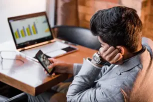 A man sitting at a desk with a laptop in front of him, casually browsing his phone.